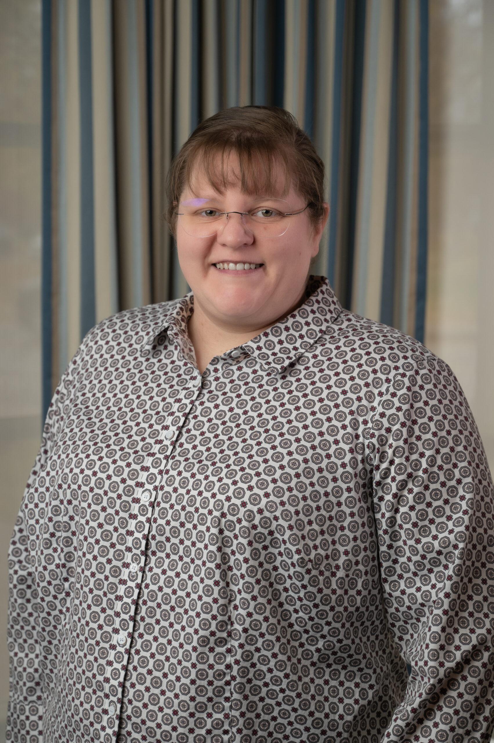 Megan Torres stands in front of a gray background. She wears glasses and is smiling. She is wearing a black and white button down